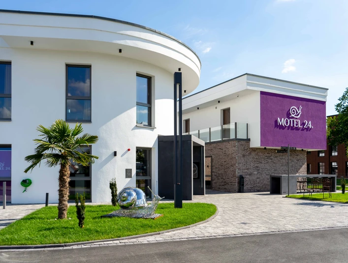 Modernes Motelgebäude mit weißer Fassade und violettem Motel 24h-Logo, umgeben von Rasenflächen.
