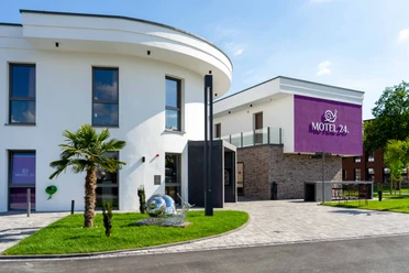 Motel24-Außenansicht Modernes Motelgebäude mit weißer Fassade und violettem Motel 24h-Logo, umgeben von Rasenflächen.