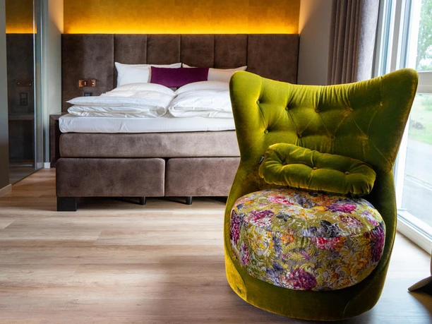 Gemütliches Hotelzimmer mit modernem Bett, grünem Sessel und bunter Kissen, helle Atmosphäre.