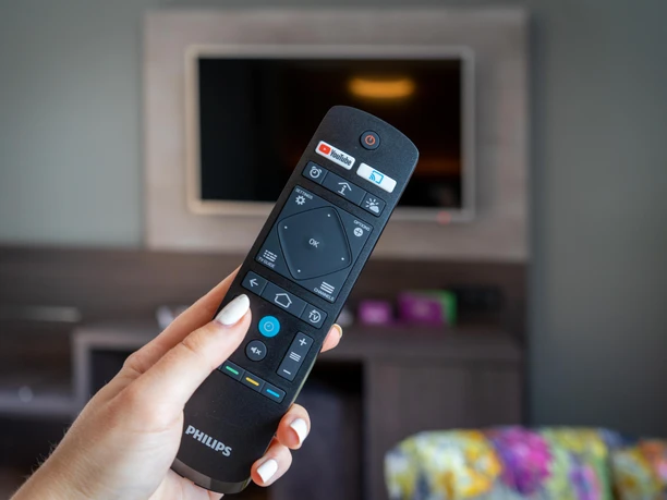 Eine Hand hält eine TV-Fernbedienung von Philips, im Hintergrund ein unscharfer Fernseher.