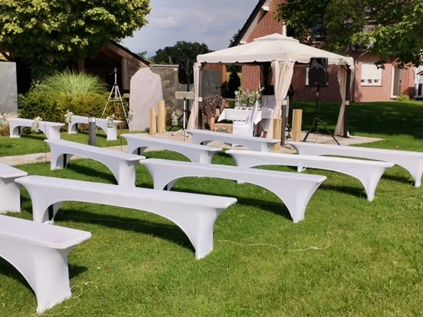 Weiße, festlich dekorierte Bänke im Freien, ausgerichtet auf einen überdachten Altar für eine Zeremonie.