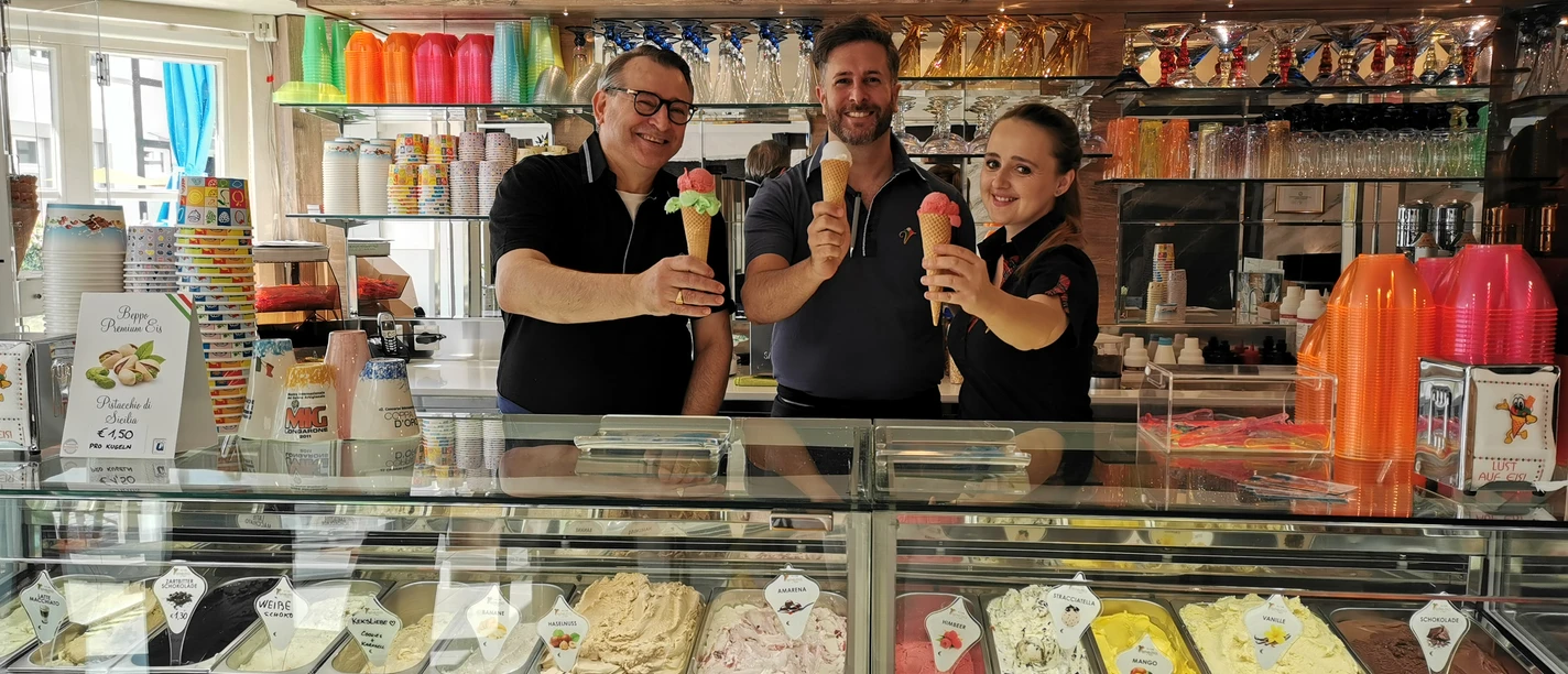 Eiscafé Venezia_Familie Schwarz Drei Personen präsentieren stolz bunte Eiscremekugeln hinter einer Vitrine mit vielfältigen Eissorten.