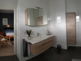 Ein modernes Badezimmer mit hölzernen Schränken, großer Spiegel, Waschbecken und Blick in ein Schlafzimmer.