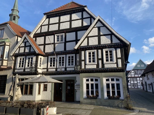 1643_Außenansicht_02 Fachwerkhaus mit weißen Wänden und schwarzen Holzbalken, daneben gotischer Kirchturm unter blauem Himmel.