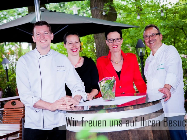 Vier lächelnde Personen in schicker Kleidung stehen um einen Tisch in einem grünen Gartenbereich.