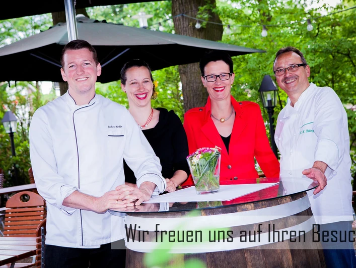 Vier lächelnde Personen in schicker Kleidung stehen um einen Tisch in einem grünen Gartenbereich.