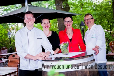 Domschenke_Familie Vier lächelnde Personen in schicker Kleidung stehen um einen Tisch in einem grünen Gartenbereich.