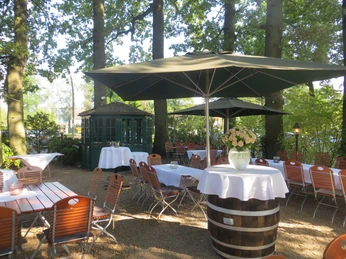 Schattiger Biergarten mit Holzmöbeln und Sonnenschirmen, umgeben von hohen Bäumen und grüner Natur.