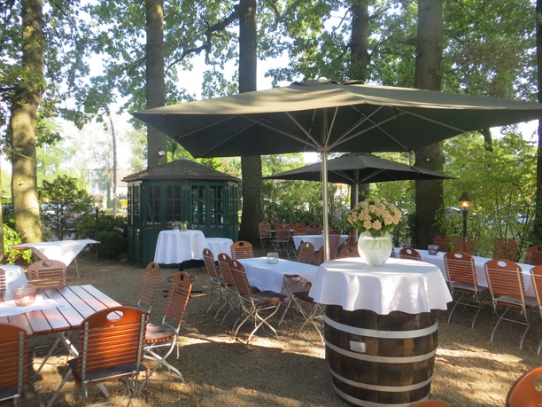 Schattiger Biergarten mit Holzmöbeln und Sonnenschirmen, umgeben von hohen Bäumen und grüner Natur.