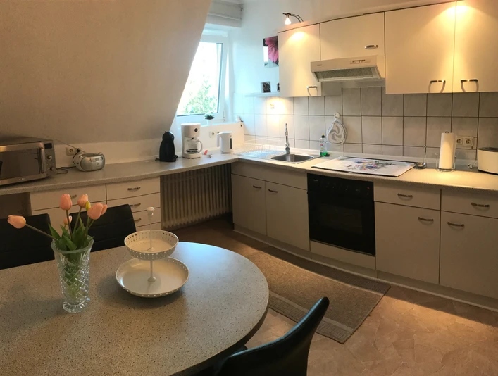 Gartenblick (1).jpeg Helle Küche mit Fenster, Herd, Mikrowelle, Kaffeemaschine, Esstisch und Blumen in einer Vase.