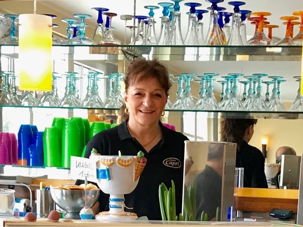 Eine lächelnde Frau hinter einer Bar, umgeben von bunten Gläsern und Dekorationen.