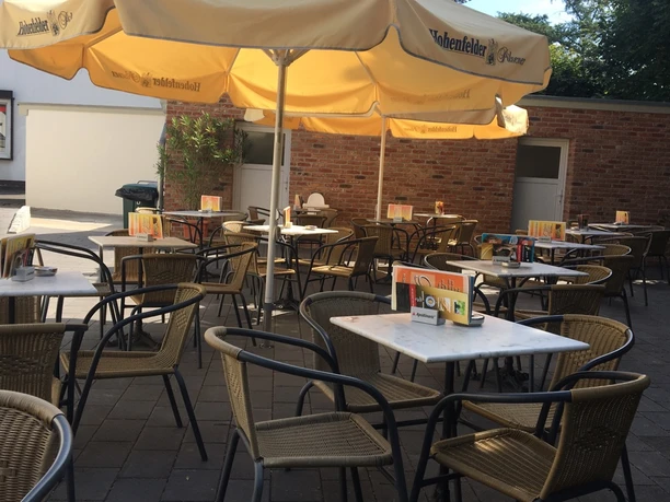 Gemütlicher, sonniger Biergarten mit gelben Schirmen, leeren Tischen und Stühlen auf gepflastertem Boden.