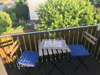 Holzveranda mit zwei Stühlen, blauem Kissen, kleinem Tisch und Pflanze, umgeben von grünem Garten.