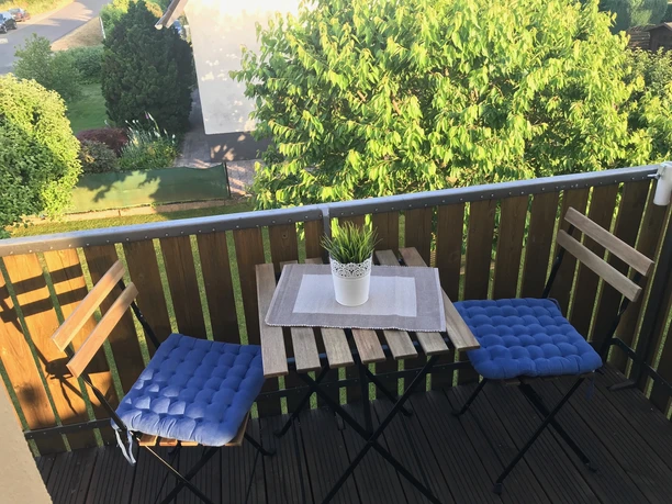 Holzveranda mit zwei Stühlen, blauem Kissen, kleinem Tisch und Pflanze, umgeben von grünem Garten.