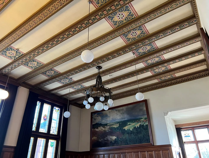 Ratssaal Rathaus Hoya Holzvertäfelter Ratssaal mit prunkvollem Kronleuchter, gemusterten Deckenbalken und bemaltem Wandbild.