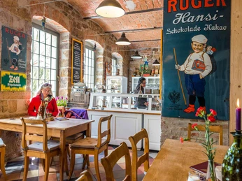 Gemütliches Café mit Holztischen, einer Frau in roter Jacke, Vintage-Werbeplakaten an den Wänden und einer Vitrine mit Kuchen.A cozy café with wooden tables, a woman in a red jacket, vintage advertising posters on the walls and a display case with cakes.Útulná kavárna s dřevěnými stolky, ženou v červeném saku, historickými reklamními plakáty na stěnách a vitrínou s dorty.Przytulna kawiarnia z drewnianymi stolikami, kobietą w czerwonej kurtce, plakatami reklamowymi w stylu vintage na ścianach i gablotą z ciastami.Gezellig café met houten tafels, een vrouw in een rood jasje, vintage reclameposters aan de muur en een vitrine met gebak.Accogliente caffetteria con tavoli in legno, una donna in giacca rossa, manifesti pubblicitari d'epoca alle pareti e una vetrina con torte.