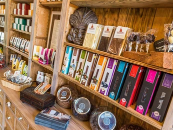 Adoratio Schokoladenkunst Regale mit bunter Schokoladenverpackung, Schokoladenlollis und Körben in einem Holzladen; warme, einladende Atmosphäre.Shelves with colorful chocolate packaging, chocolate lollipops and baskets in a wooden store; warm, inviting atmosphere.Regály s barevnými obaly na čokoládu, čokoládová lízátka a košíky v dřevěném obchodě; příjemná, hřejivá atmosféra.Półki z kolorowymi opakowaniami czekolady, czekoladowe lizaki i kosze w drewnianym sklepie; ciepła, zachęcająca atmosfera.Schappen met kleurrijke chocoladeverpakkingen, chocoladelolly's en manden in een houten winkel; warme, uitnodigende sfeer.Scaffali con confezioni di cioccolato colorate, lecca-lecca e cestini di cioccolato in un negozio di legno; atmosfera calda e invitante.