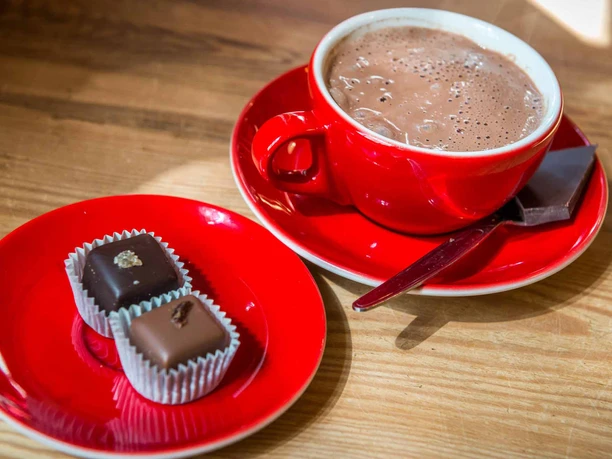 Adoratio Schokoladencafé Heiße Schokolade & Pralinen Rote Tasse mit heißer Schokolade auf Untertasse, daneben ein roter Teller mit zwei Pralinen auf hellem Holztisch.