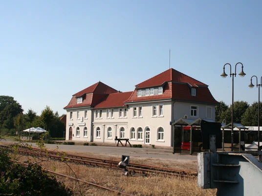 Bahnhof Hoya in Niedersachsen: Historisches Gebäude mit rotem Dach und weißen Fassaden, Gleise im Vordergrund.