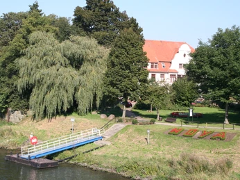 Rathaus Hoya Rotes Ziegeldach des Rathauses Hoya hinter Bäumen, Wiese mit "Hoya" Blumenbeet am Flussufer.