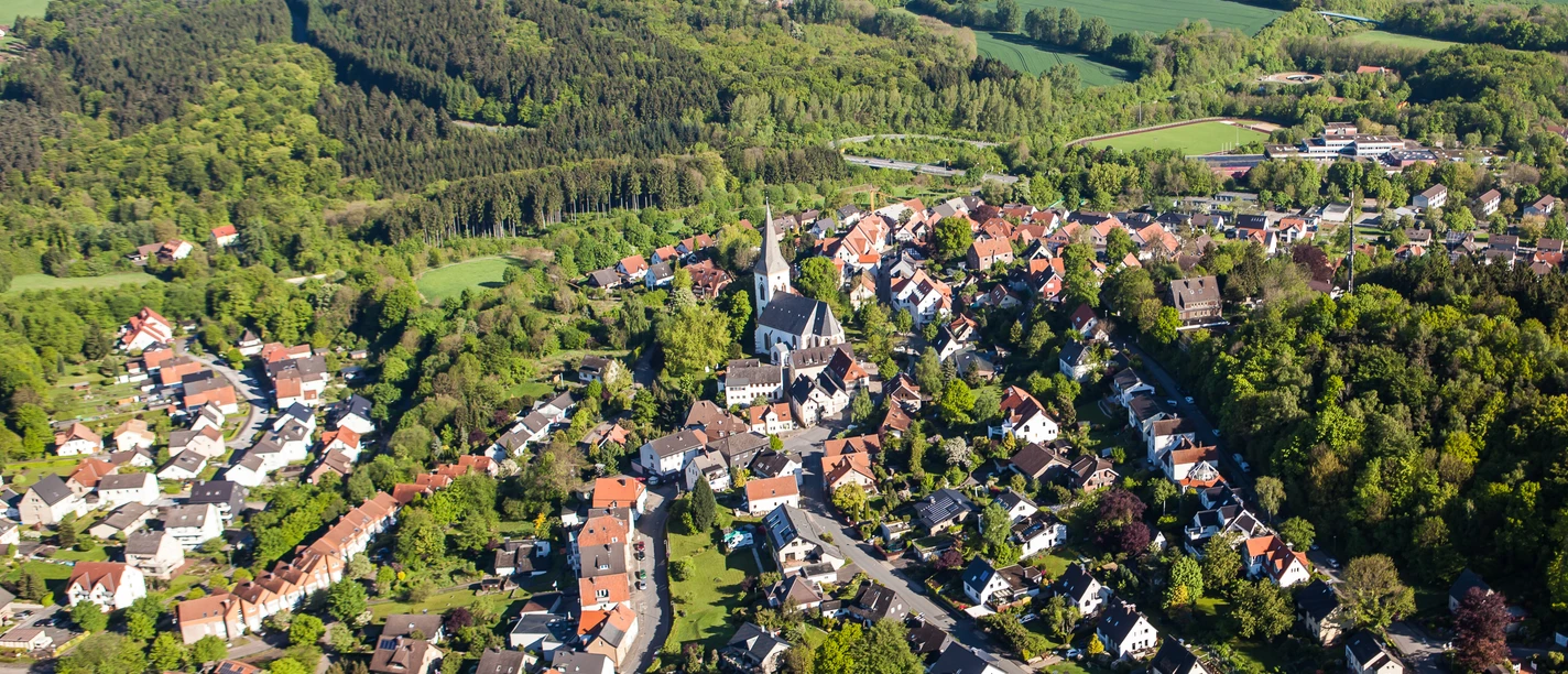 Stadt Oerlinghausen – Fotograf Patrick Piecha Luftaufnahme einer grünen Hügelstadt mit vielen Häusern, einer Kirche und umgeben von Wäldern.