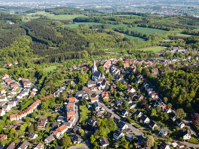 Stadt Oerlinghausen – Fotograf Patrick Piecha Luftaufnahme einer grünen Hügelstadt mit vielen Häusern, einer Kirche und umgeben von Wäldern.
