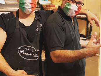 Zwei Männer in einer Pizzeria tragen italienische Gesichtsmasken und posieren fröhlich in der Küche.