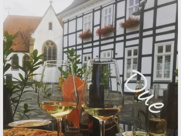 Zwei Gläser Weißwein auf einem Tisch vor einem Fachwerkhaus mit weißen Wänden und blumengeschmückten Fenstern.
