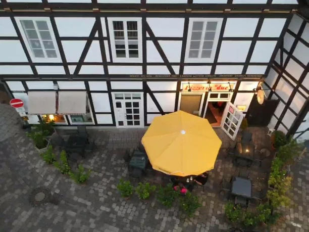 Fachwerkhaus mit gelbem Sonnenschirm vor einem Café, umgeben von gepflasterter Straße und Pflanzen.