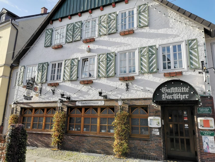 Gaststätte Bartscher_Außenansicht Traditionelles Fachwerkgebäude mit grünen Fensterläden, darunter das Schild "Gaststätte Bäcker".
