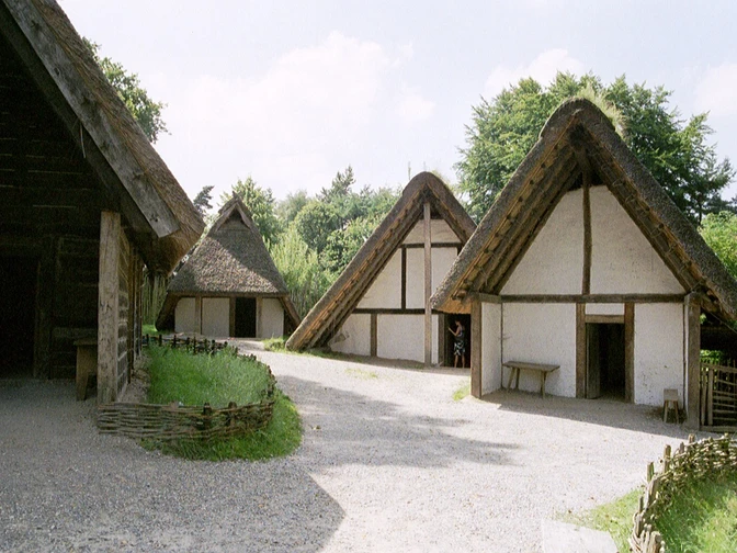AFM_Große_Bergstadt_Runde_1.JPG Drei rekonstruierte historische Fachwerkhäuser mit Strohdächern in einem Freilichtmuseum bei Sonnenschein.