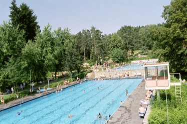 Freibad 1999 zwei Becken_GroßeBergstadtrunde_2.JPG Zwei große Schwimmbecken im Freibad von Bäumen und Liegewiese umgeben; Menschen entspannen.