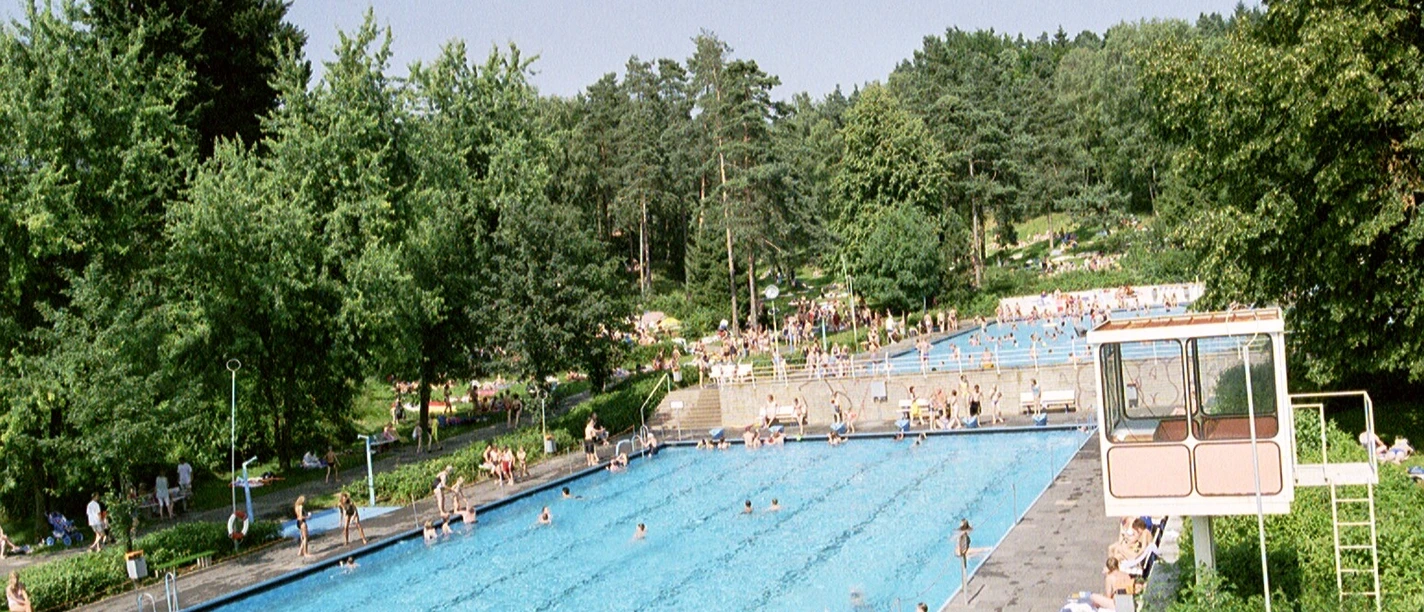 Freibad 1999 zwei Becken_GroßeBergstadtrunde_2.JPG Zwei große Schwimmbecken im Freibad von Bäumen und Liegewiese umgeben; Menschen entspannen.