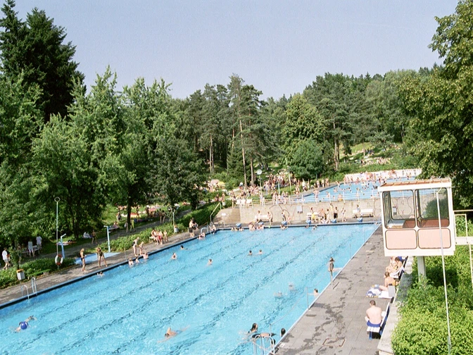 Freibad 1999 zwei Becken_GroßeBergstadtrunde_2.JPG Zwei große Schwimmbecken im Freibad von Bäumen und Liegewiese umgeben; Menschen entspannen.
