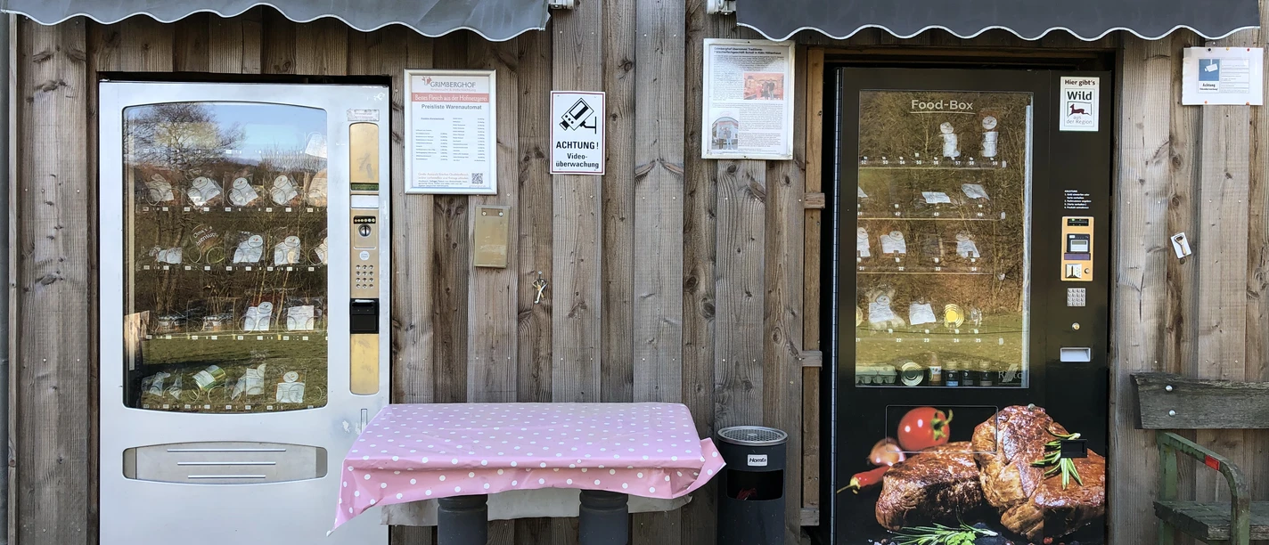 Fleischautomat am Grimberghof Zwei Automaten an einer Holzwand bieten verschiedene Waren, einer mit Fleischprodukten und außen ein Tisch.