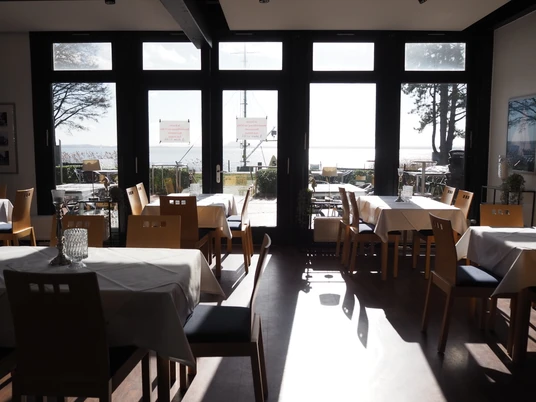 Ein lichtdurchfluteter Raum mit gedeckten Tischen und Blick durch große Fenster auf die sonnige Küste.