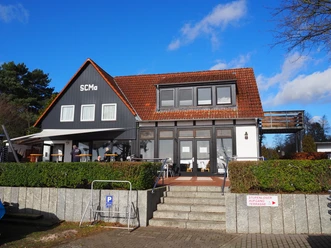A two-storey gray building with red roof tiles and a terrace under a blue sky.