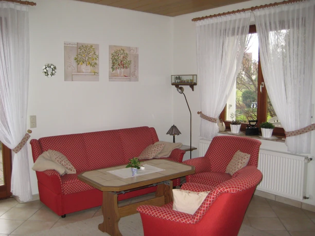 Ferienwohnung 1.jpg Wohnzimmer mit roten Sofas, Holztisch, weißer Wanddekoration und großen Fenstern mit Vorhängen.