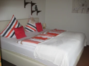 Ferienwohnung 1 Schlafzimmer Doppelbett mit gestreiften Kissen und roten Akzenten, dekorative Regale an weißer Wand.