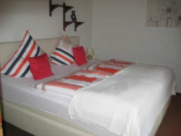 Ferienwohnung 1 Schlafzimmer Doppelbett mit gestreiften Kissen und roten Akzenten, dekorative Regale an weißer Wand.