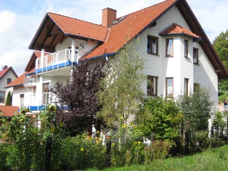 Ein großes, weißes Haus mit roten Dachziegeln, umgeben von grünen Pflanzen und Bäumen unter klarem Himmel.
