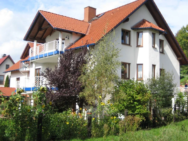 Ein großes, weißes Haus mit roten Dachziegeln, umgeben von grünen Pflanzen und Bäumen unter klarem Himmel.