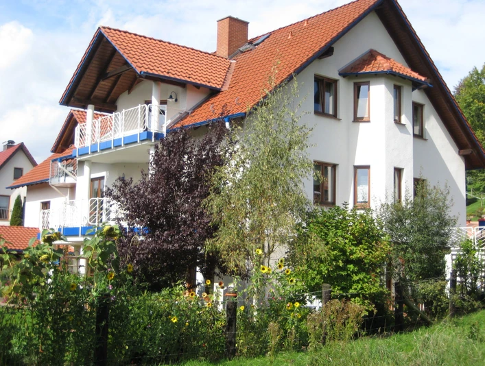 FEWOSilbermann.jpg Ein großes, weißes Haus mit roten Dachziegeln, umgeben von grünen Pflanzen und Bäumen unter klarem Himmel.