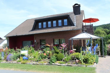 Haus Heidegarten.jpg Zweistöckiges Backsteinhaus mit gepflegtem, blühendem Garten und einem markanten Pavillon davor.