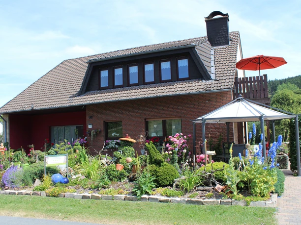 Haus Heidegarten.jpg Zweistöckiges Backsteinhaus mit gepflegtem, blühendem Garten und einem markanten Pavillon davor.