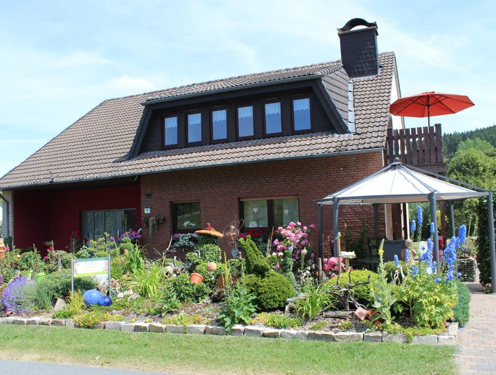Haus Heidegarten.jpg Zweistöckiges Backsteinhaus mit gepflegtem, blühendem Garten und einem markanten Pavillon davor.