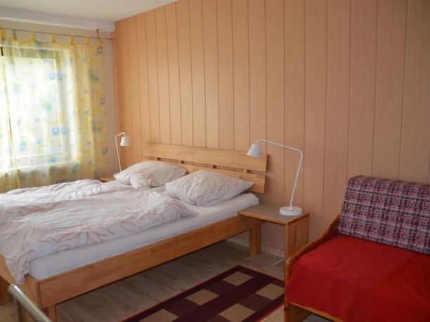 Helles Schlafzimmer mit Doppelbett und rotem Sofa, Holzwände, Tageslichtfenster mit gelben Vorhängen.