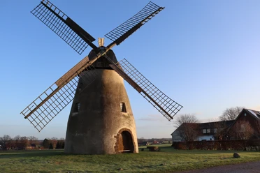 Mühle Auf der Höchte Historische Windmühle mit Holzflügeln auf einer Wiese, umgeben von Bäumen und einem blauen Himmel.