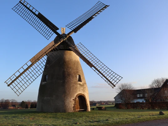 Mühle Auf der Höchte Historische Windmühle mit Holzflügeln auf einer Wiese, umgeben von Bäumen und einem blauen Himmel.