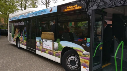 HeideShuttle Bus mit Landschaftsmotiven, Schafen und Heide beklebt. Tür offen, Fahrgäste steigen ein.